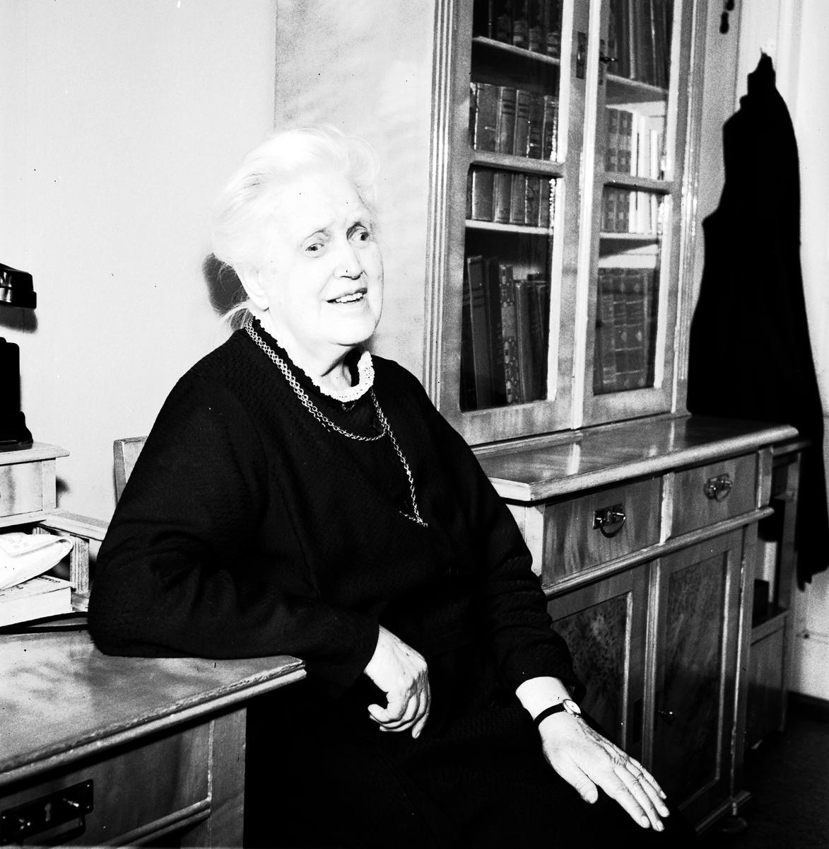 Svartvit porträttbild av en äldre kvinna med vitt hår och mörka kläder. Hon sitter avslappnat vid ett skrivbord, med en bokhylla i bakgrunden fylld med böcker. Kvinnan ler vänligt och bär ett halsband samt en klocka på vänster handled.