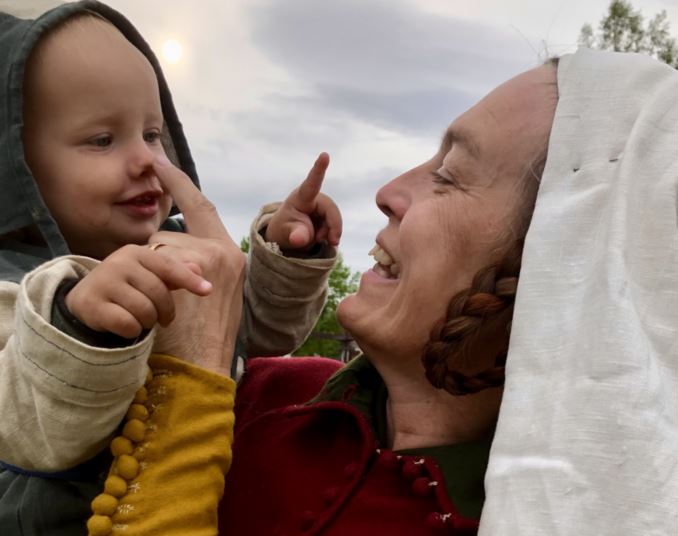 En kvinna och ett litet barn delar ett kärleksfullt ögonblick utomhus. Kvinnan, klädd i traditionella medeltidskläder med vit huvudduk, ler varmt medan hon försiktigt rör vid barnets näsa. Barnet, klädd i en grå och grön huvtröja, ler och pekar tillbaka på kvinnan med sitt finger. Solen skiner svagt genom en molnig himmel i bakgrunden.