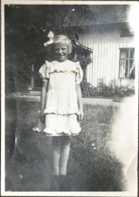 En flicka i vit klänning, vit rosett och blont hår står framför ett hus