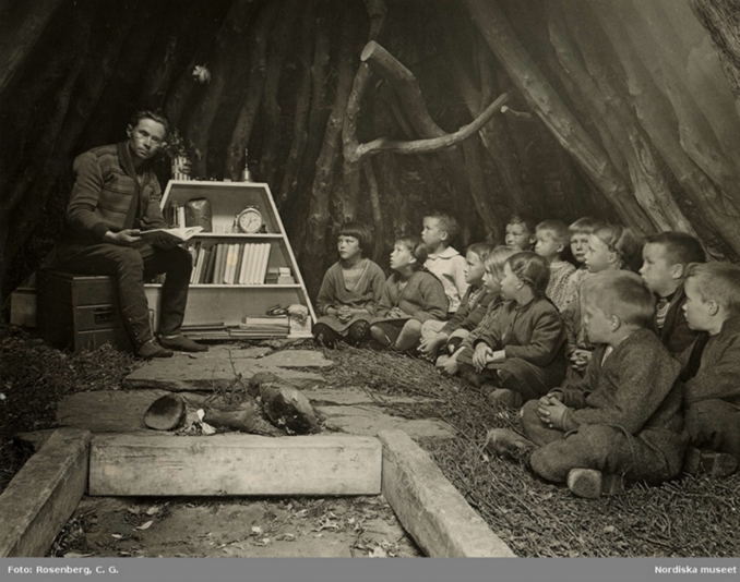 En grupp barn sitter i ett tält, en lärare sitter med en uppslagen bok.