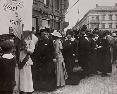 En grupp kvinnor står utomhus i en demonstration, hållandes en banderoll med texten 'Rösträtt för kvinnor' Göteborgs stadsbild syns i bakgrunden