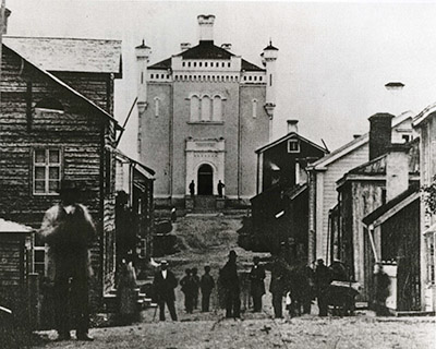 En gammal bild av en stad med människor som promenerar längs gatan. I slutet av gatan, uppe på en höjd, reser sig en stor och hög byggnad med två torn