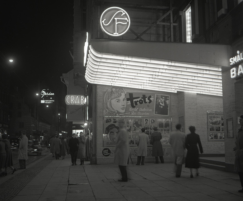 En biograf i Malmö år 1951 med neonljus och filmaffischer, där människor står utanför och tittar på affischer för filmen 'Trots'