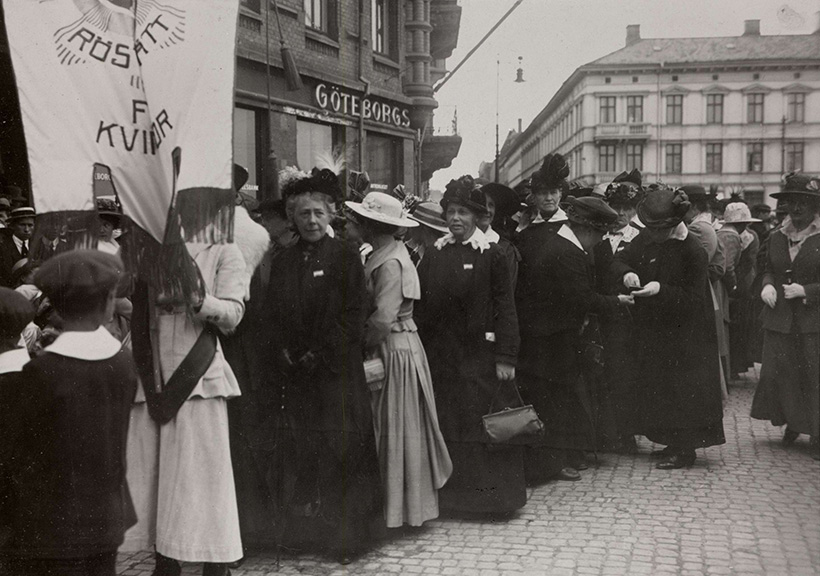 En grupp kvinnor står utomhus i en demonstration, hållandes en banderoll med texten 'Rösträtt för kvinnor.' Göteborgs stadsbild syns i bakgrunden