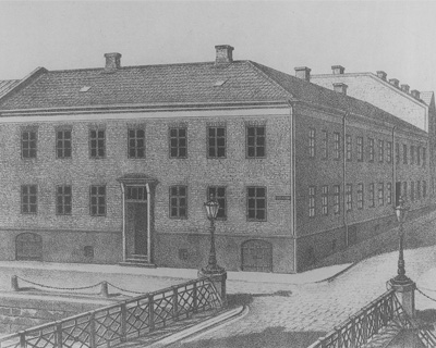 En teckning av ett stort tegelhus vid en bro över vatten, med lyktstolpar och kullerstensgata framför.