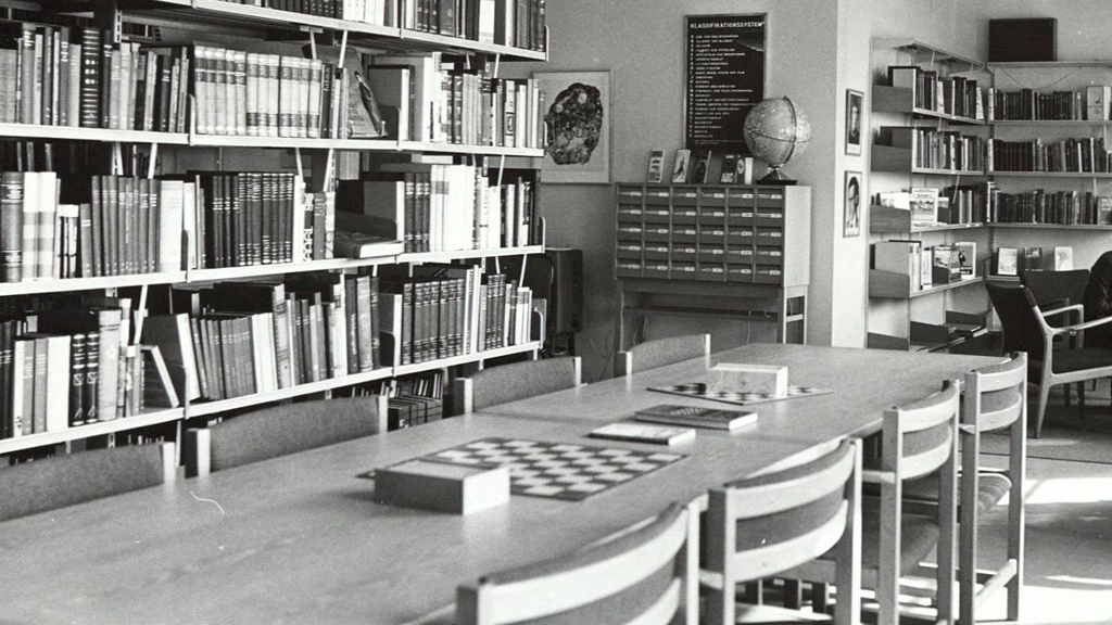 Ett bibliotek med bokhyllor, ett bord med stolar och ett schackbräde, samt en klassificeringsskylt på väggen