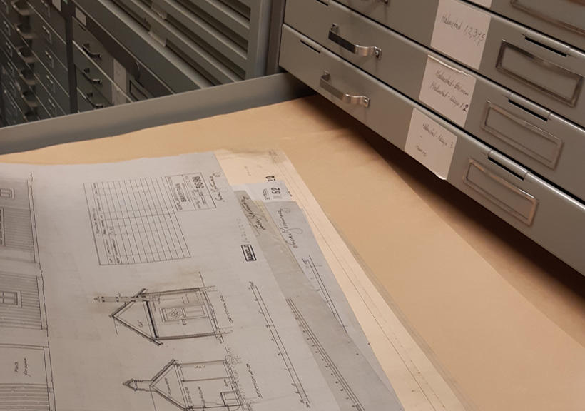 En öppen låda med ritningar i ett arkivskåp. Ritningarna visar tekniska skisser av en byggnad med detaljerad information. Arkivskåpet har flera lådor märkta med etiketter