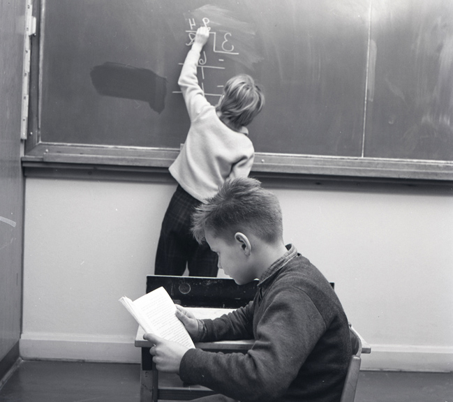 Två elever i en skolmiljö, en står vid en svart tavla och skriver, medan den andra sitter vid sin bänk och läser en bok