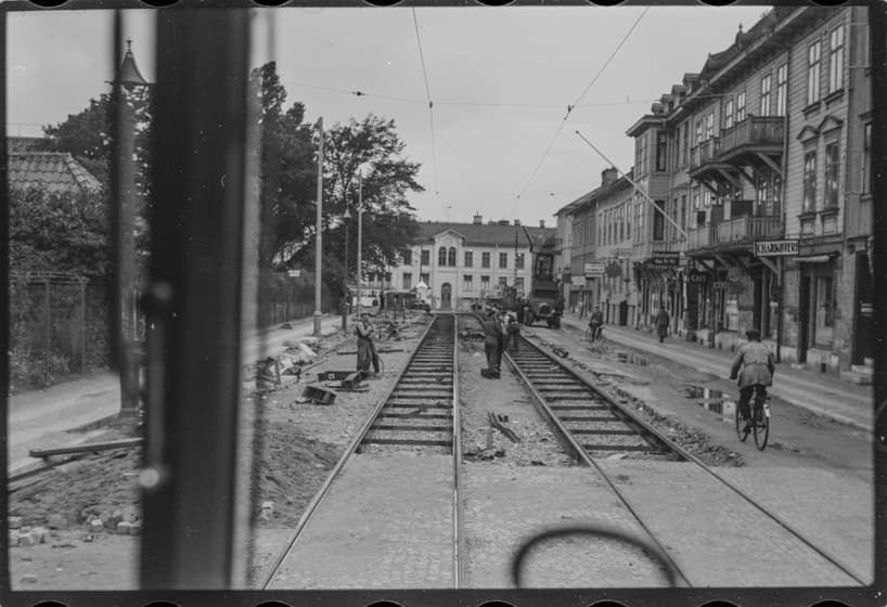 Svart/vit bild från 1938. Fotograferad från en tågräls, längs med rälsen. En bit fram svänger rälsen till vänster. Bortanför rälsen står ett ljust hus. Till höger står en rad med hus. En cyklist cyklar vid sidan av rälsen. Ytterligare fem personer vistas på och omkring rälsen. Till vänster går en väg och på andra sidan vägen kan man skymta fler hus.