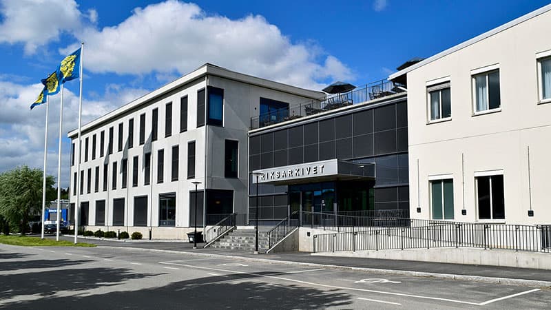 Riksarkivets moderna byggnad i Täby med vit fasad och stora fönster. Tre flaggor med arkivets emblem vajar framför byggnaden under en blå himmel.