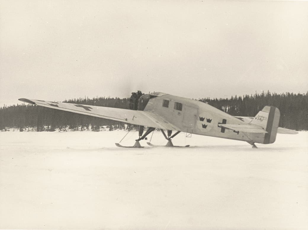 Ett svartvitt fotografi som visar ett äldre ambulansflygplan stående på snötäckt mark, utrustat med skidor för landning. Planet har svenska symboler, inklusive tre kronor, målade på sidan och bakpartiet. En person är synlig vid flygplansdörren, och i bakgrunden skymtar en tät skog. Bilden förmedlar en historisk känsla av tidigt flyg inom vinterlandskap.