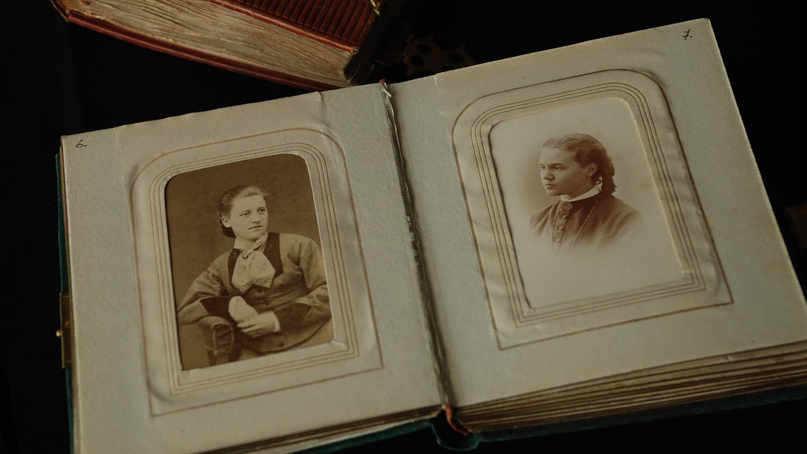 En uppslagen gammal fotoalbum med två sepiafärgade porträttbilder av unga kvinnor, placerade i dekorativa ramutskärningar på vardera sida av uppslaget. Albumets sidor är slitna, vilket förstärker känslan av dess ålder och historiska värde. I bakgrunden syns en del av en annan bok.