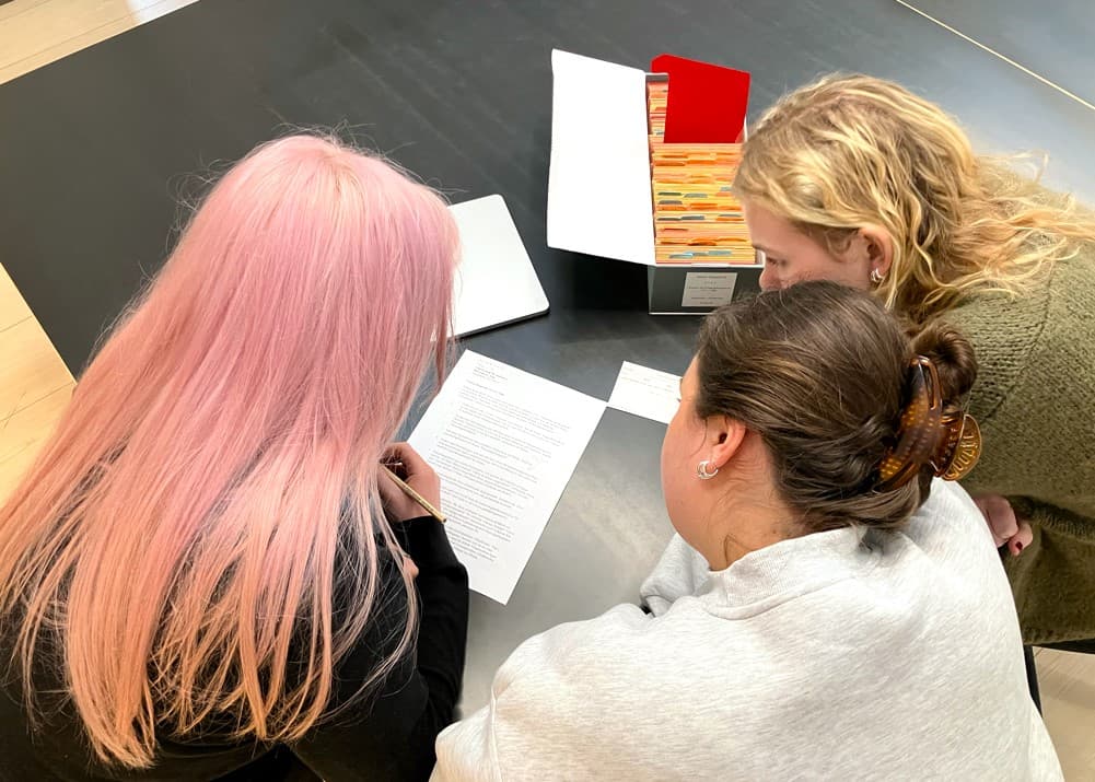 Tre personer sitter vid ett bord och granskar ett dokument tillsammans. Bakom dem finns en öppen arkivlåda med färgade flikar.