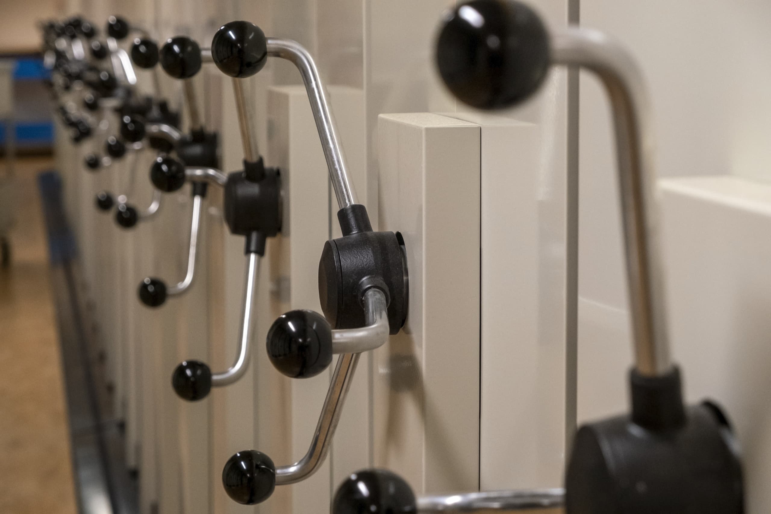 Fotografi av ett arkiv- eller magasinsutrymme med flera vita, mobila hyllsektioner. Varje sektion har en stor handvev med svarta grepp, placerad i rad längs hyllornas front. Miljön är funktionell och ordnad, med fokus på effektiv förvaring. Färgskalan är neutral med inslag av vitt, grått och svart. Intrycket är praktiskt och systematiskt.