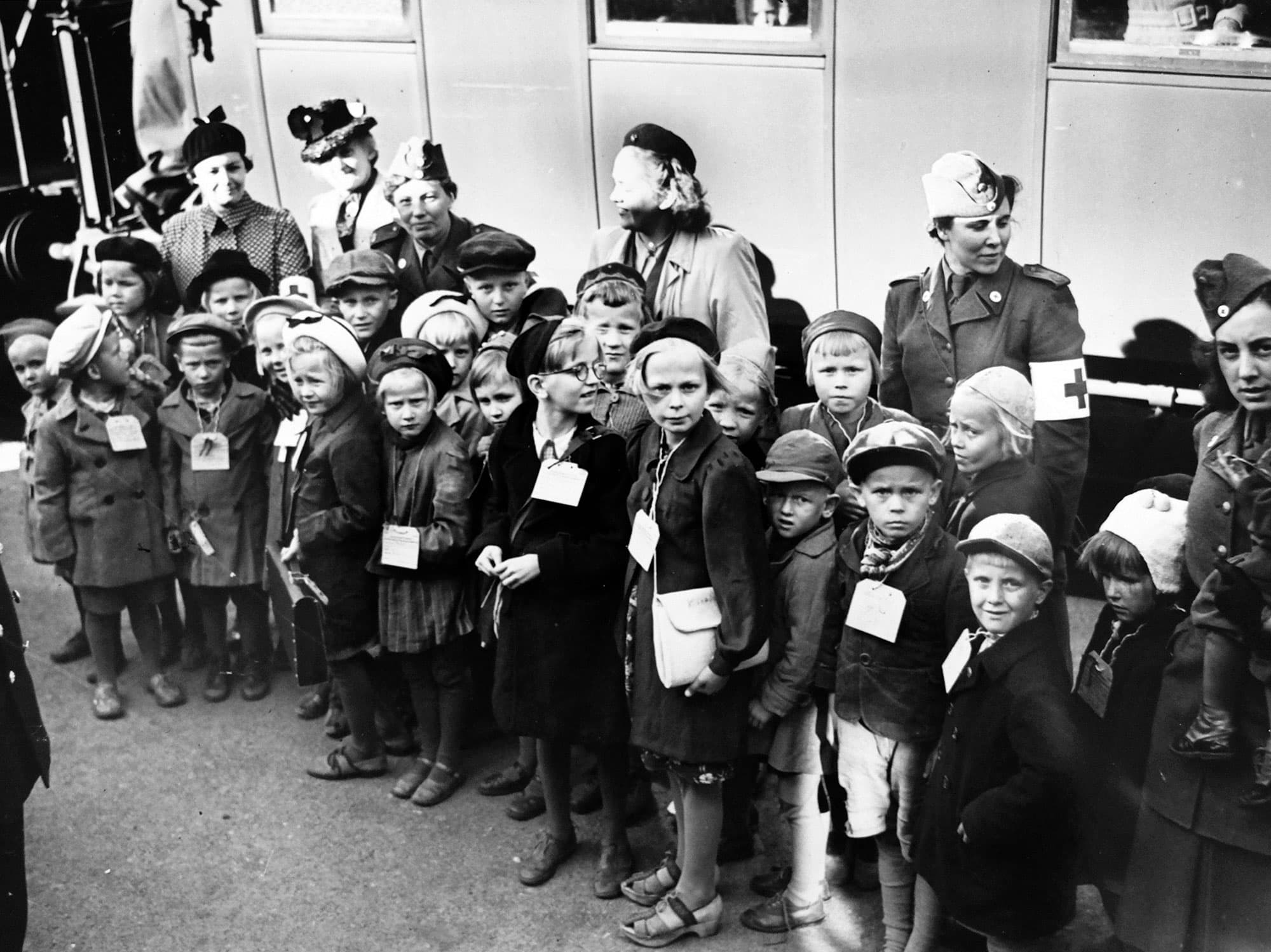 Barn i kö med Röda Kors-personal, alla bär ID-taggar, utanför tåg eller buss.