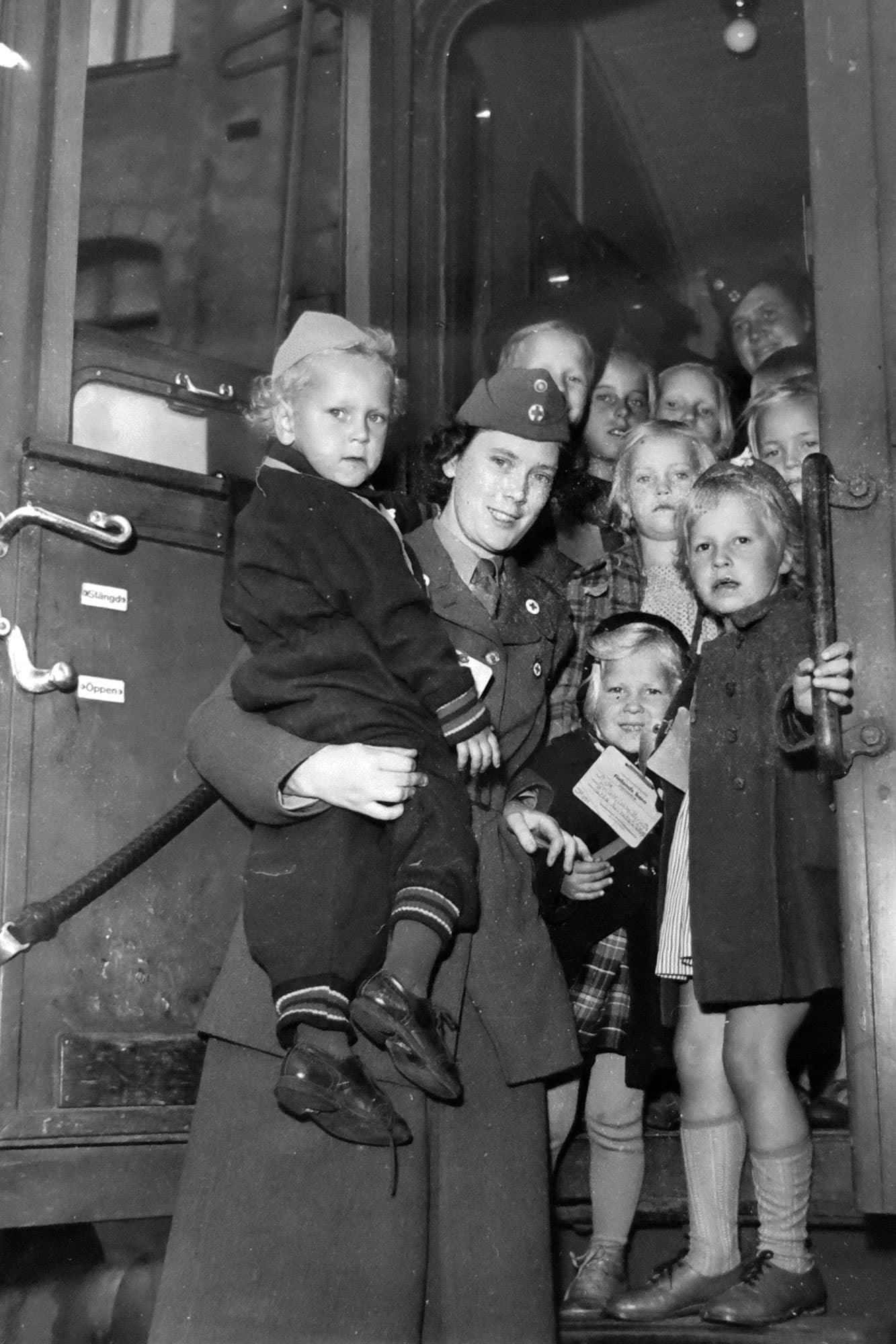 Svartvit bild: kvinna i uniform lyfter barn vid tågdörr, flera barn med ID-taggar.
