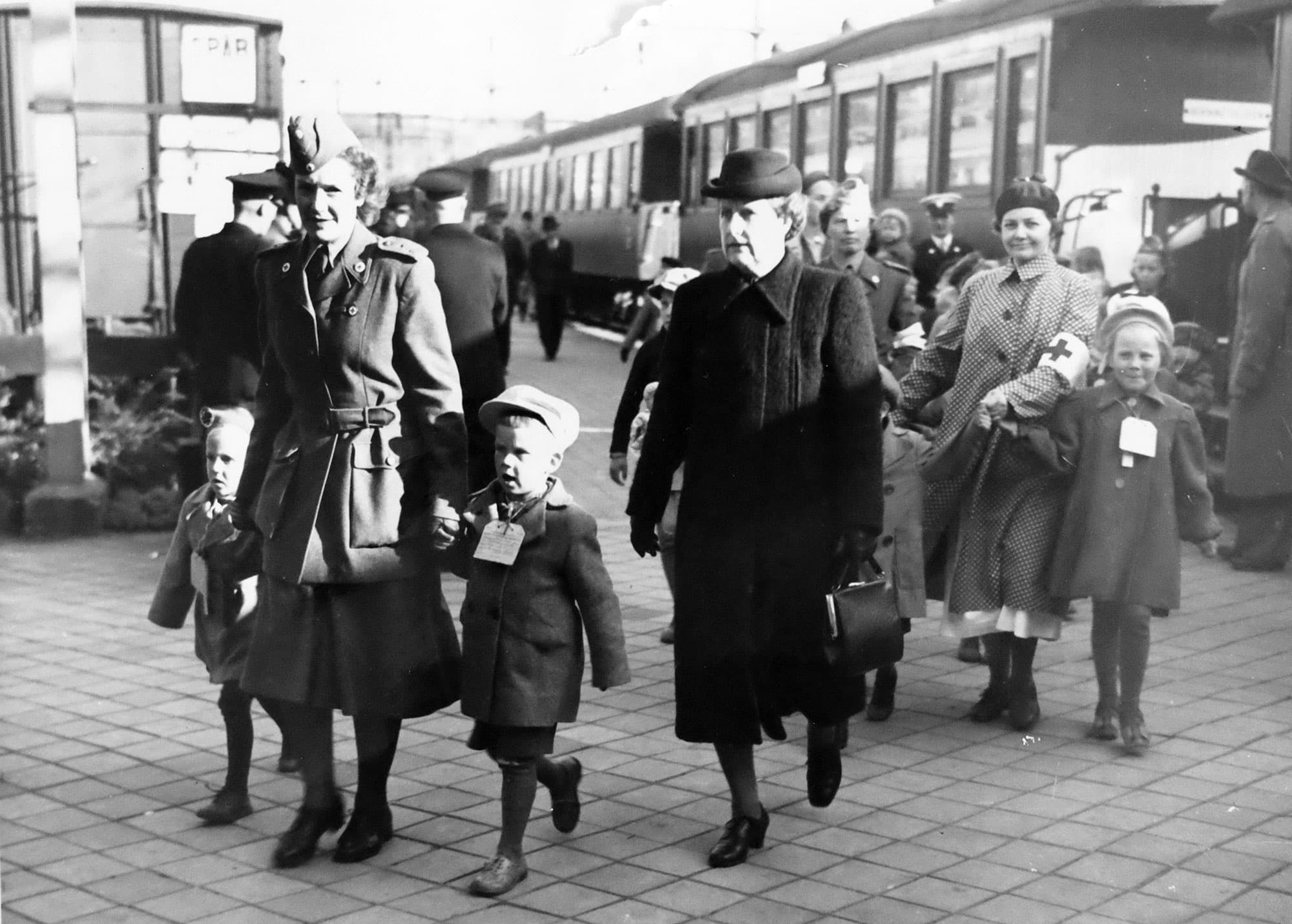 Svartvit bild på kvinnor och barn med ID-taggar på perrong, framför ett tåg.