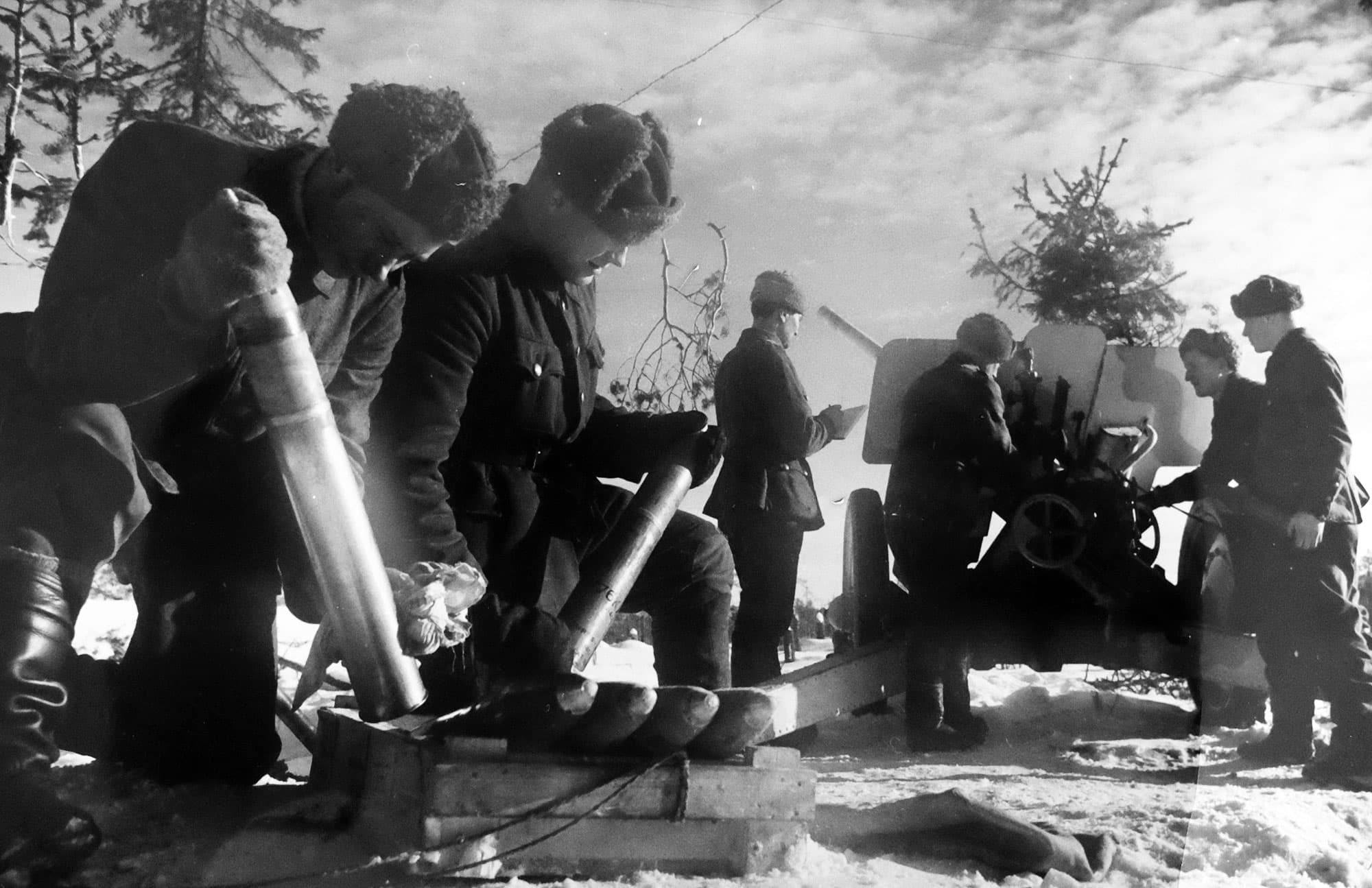 Soldater i vinteruniform laddar artilleri i snölandskap, granater ligger uppradade på träplattform.