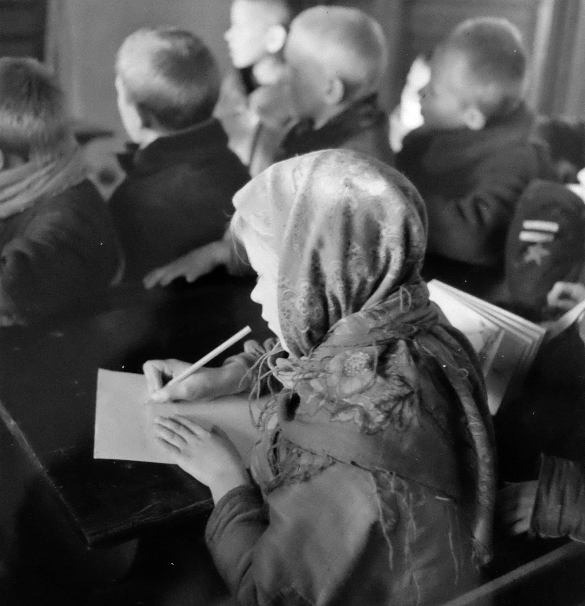 Svartvit bild på barn i klassrum, varmt klädda, ett skriver med blyertspenna.