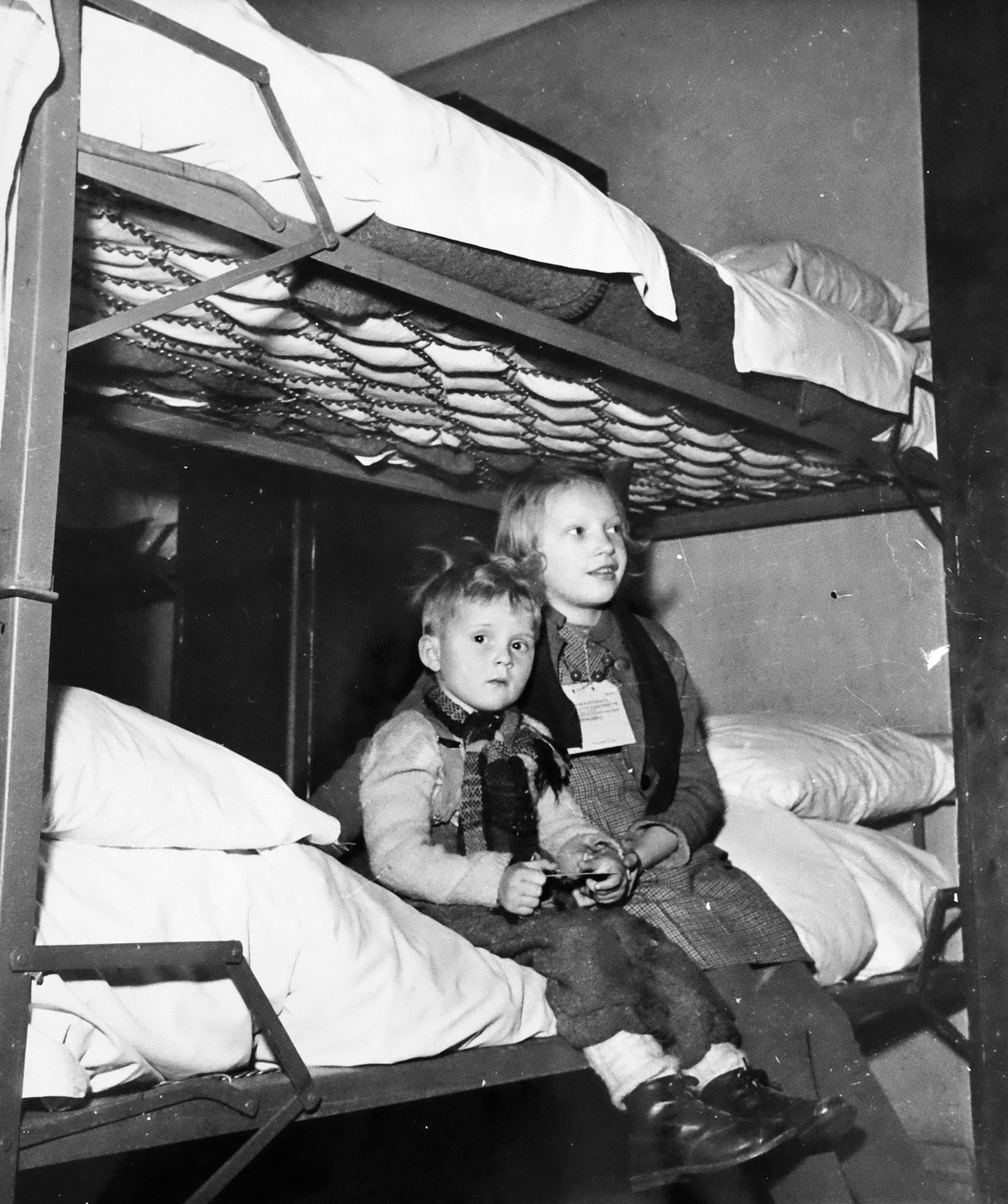 Två barn sitter på en metallvåningssäng i enkel miljö, ett bär namnbricka.