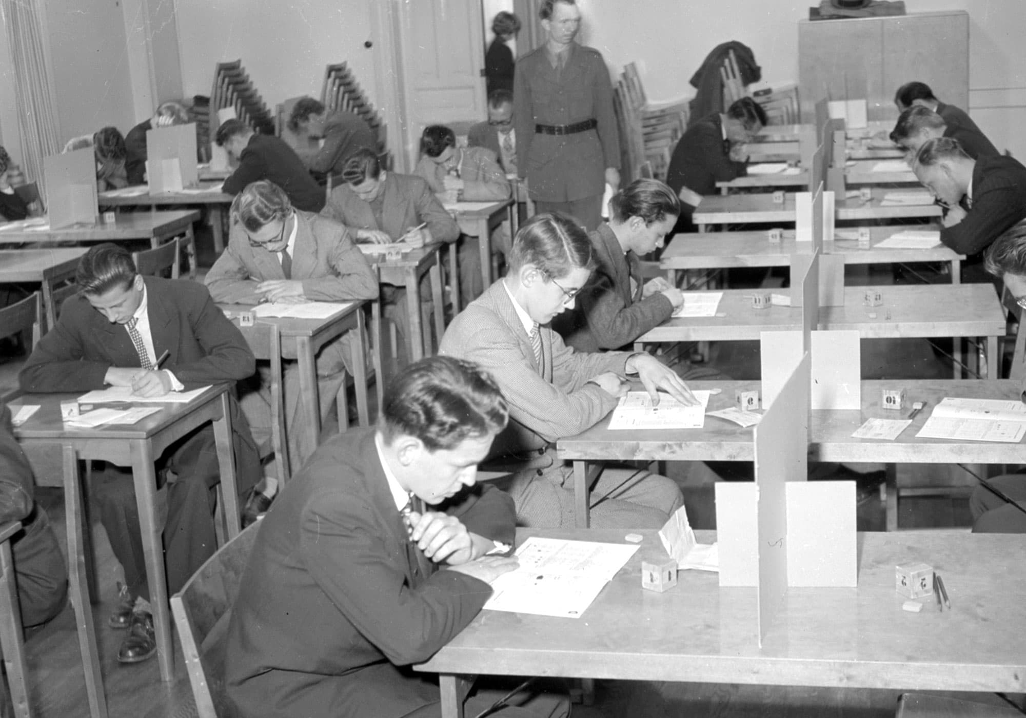 Svartvit bild visar unga män i kostym som skriver prov vid avskärmade bänkar i en övervakad skrivsal.