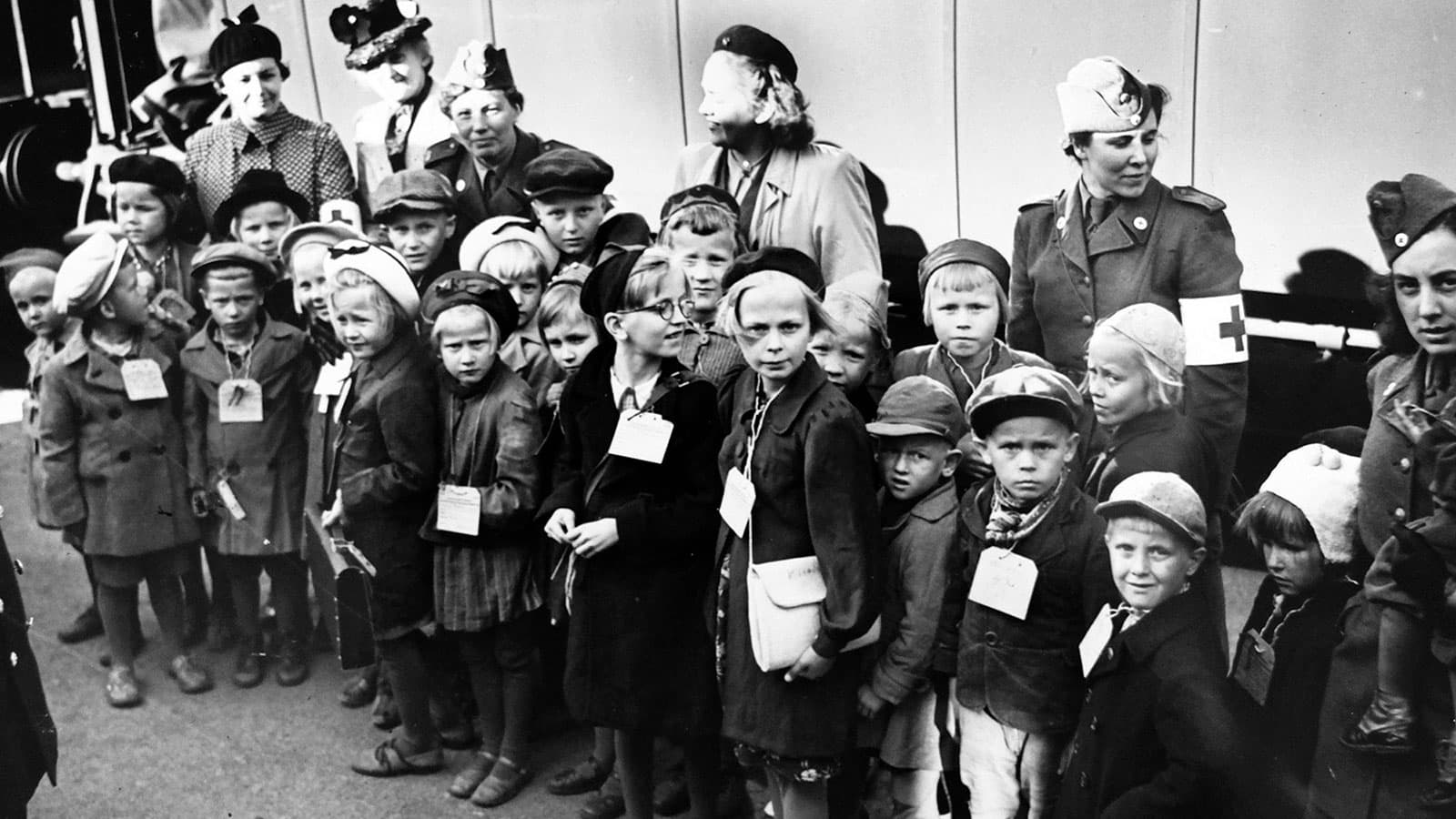 Barn och vuxna står samlade vid ett fordon. Barnen bär identifikationslappar, och några vuxna har Röda Korset-armbindlar.