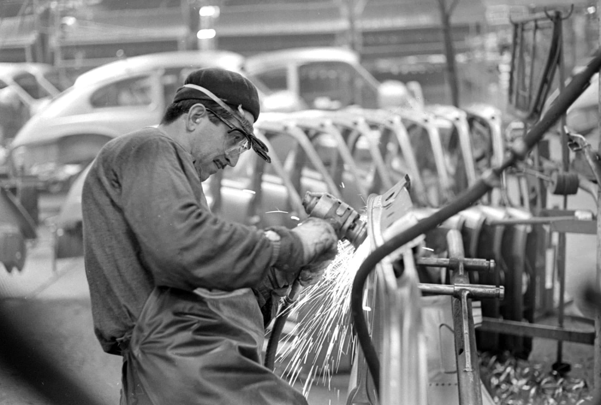 Man med skyddsglasögon svetsar i fabrik, gnistor flyger.