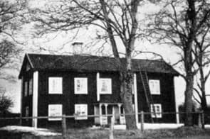 Svartvitt fotografi av hus med vita knutar och fönsterkarmar. Framför huset syns kala träd.