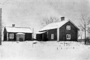 Svartvitt fotografi av två hus med vita knutar på en snötäckt mark. I bakgrunden syns kala träd.