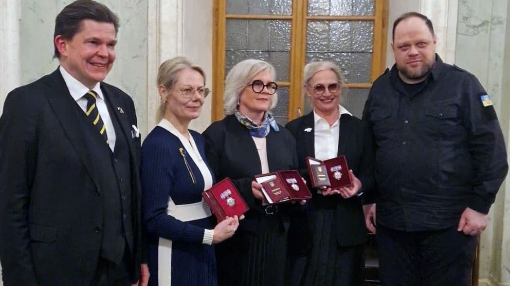 Fem personer står på rad. De tre som står i mitten håller upp varsin röd ask med medalj.