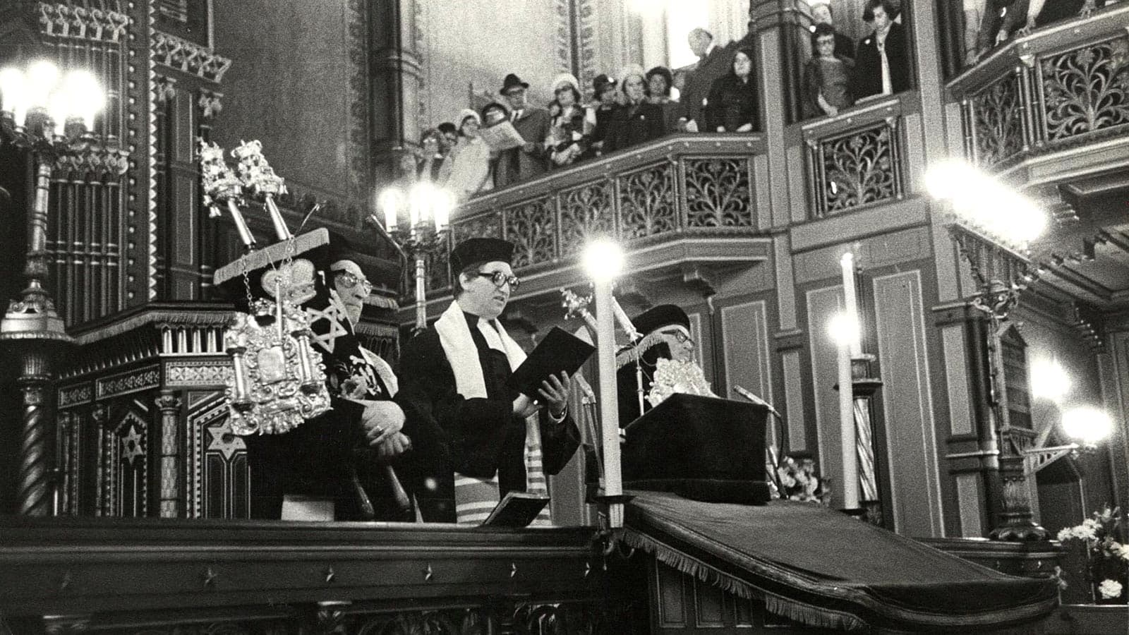 Svartvitt fotografi av en religiös ceremoni i en synagoga. Flera personer i traditionella judiska kläder står vid en upphöjd plats med Torahrullar. Rummet är rikt dekorerat och en balkong med åskådare syns ovanför.