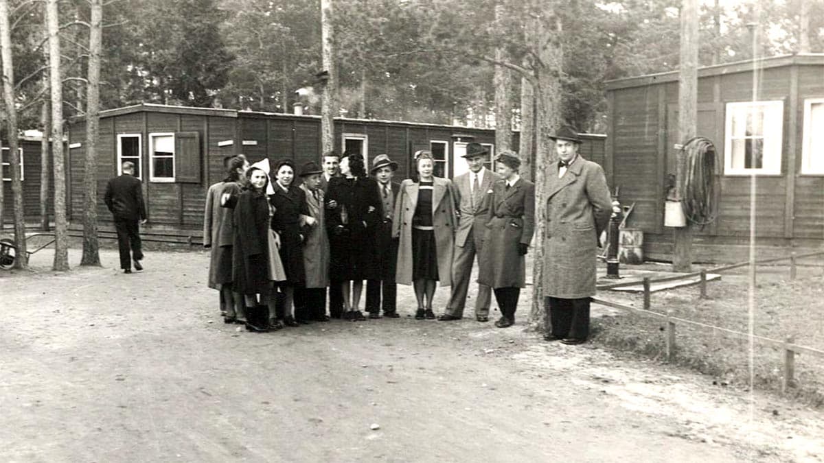 Svartvitt fotografi med människor i 1940‑talskläder som står på en grusväg mellan trähus och träd. Miljön liknar ett läger eller arbetsplats utomhus, med formell stämning och dokumentär karaktär.