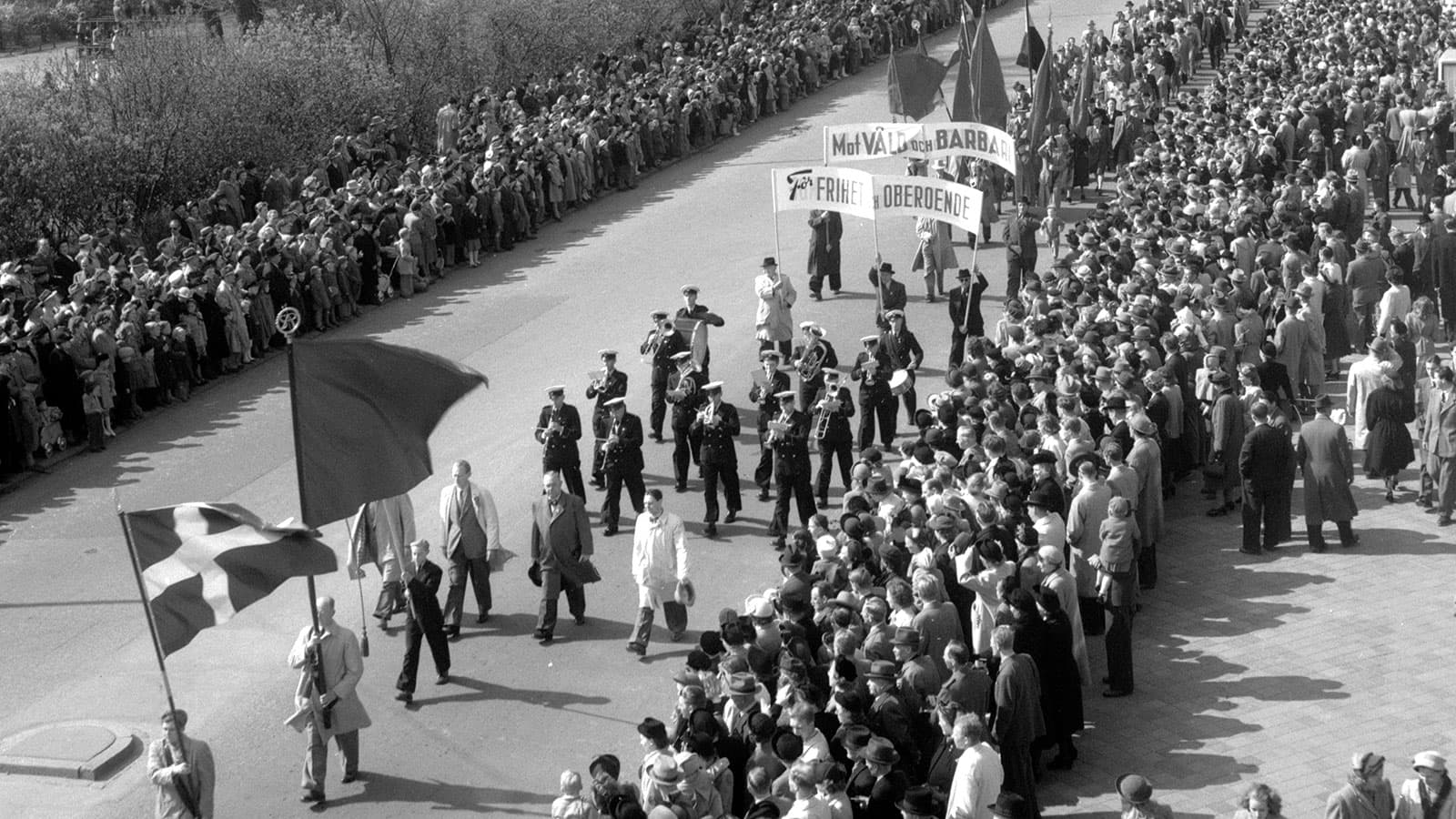 Ett svartvitt fotografi som visar ett demonstrationståg på en stadsgata. Längst fram marscherar ett uniformerat musikkår, följt av personer som bär stora fanor med texter som ”Mot våld och barbari” och ”Frihet och oberoende”. Publik står tätt längs båda trottoarerna.