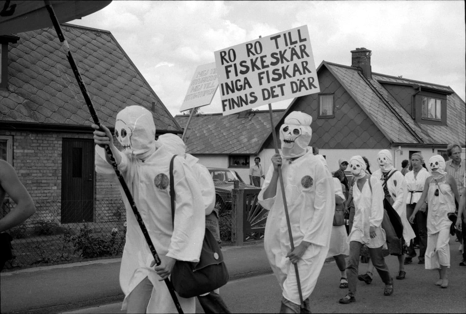 Svartvitt fotografi av personer i ett demonstrationståg. De har vita kläder och masker och bär på ett plakat där det står "ro ro till fiskeskär, inga fiskar finns det där".