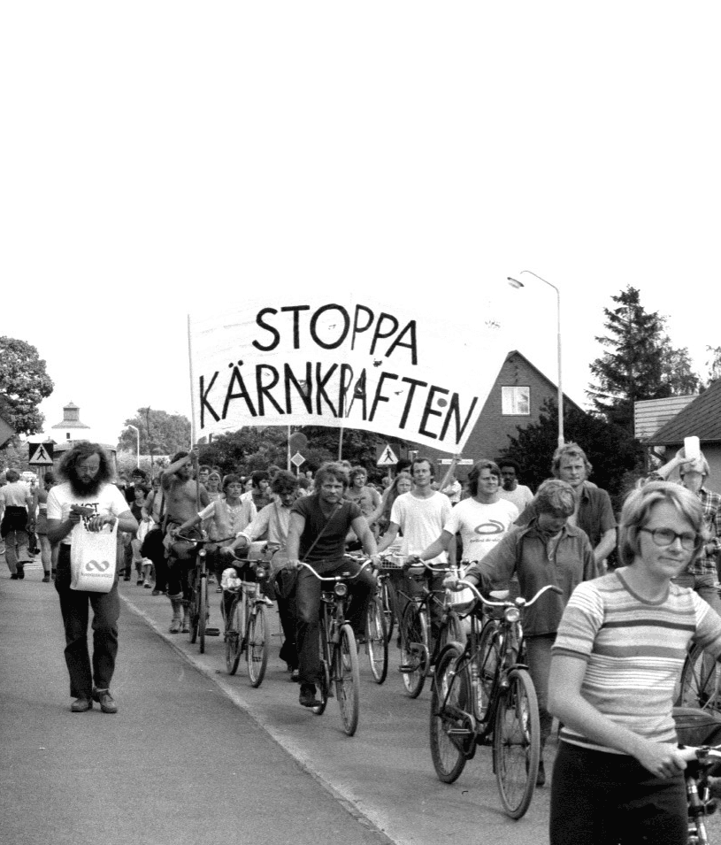 Svartvitt fotografi av personer på cyklar i ett demonstrationståg. Några bär på en stor banderoll med texten "stoppa kärnkraften".