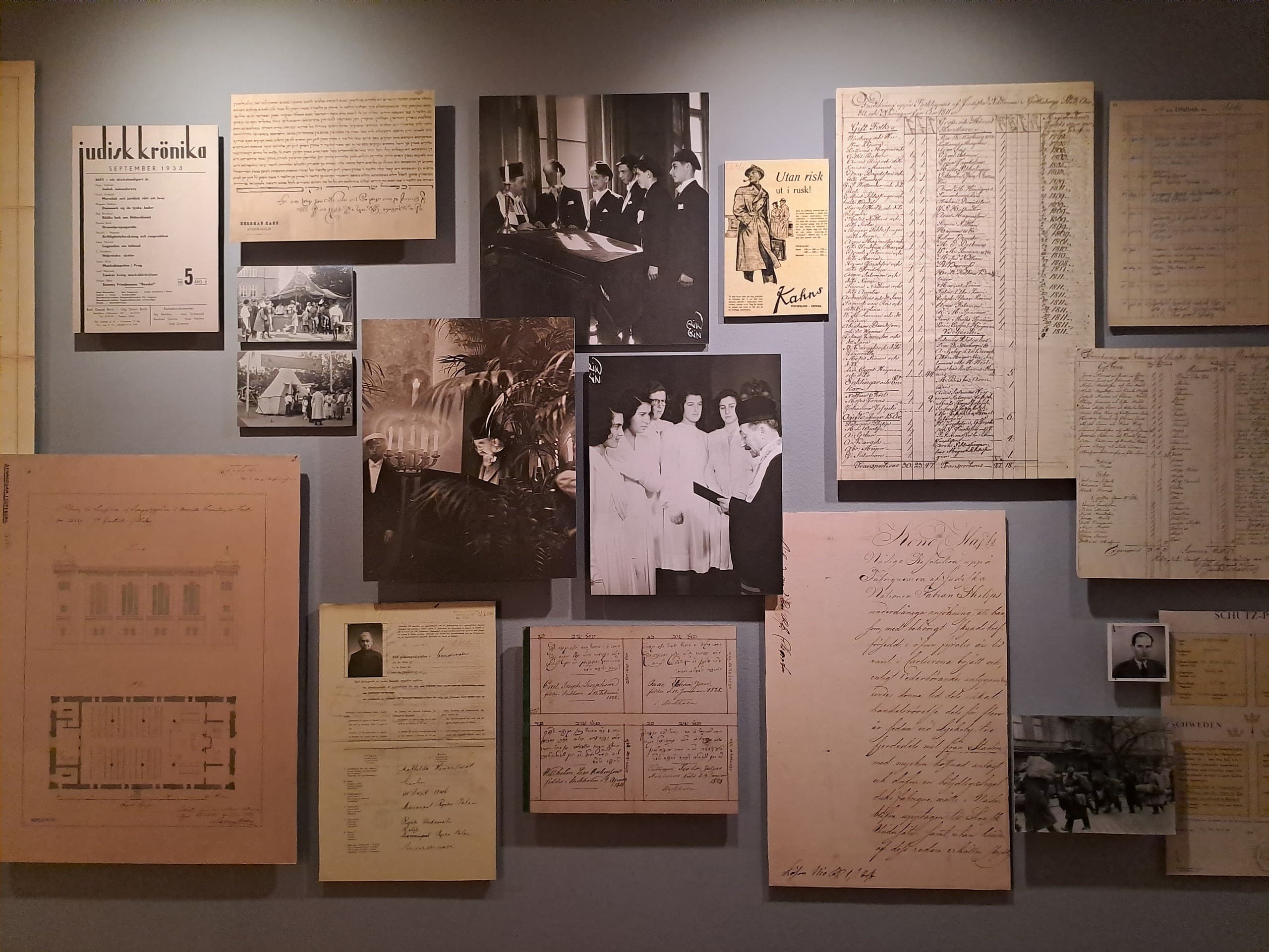 Utställningsvägg med historiska fotografier, handskrivna dokument, register, ritningar och tidningssidan Judisk krönika, som tillsammans skildrar judiskt liv i Sverige