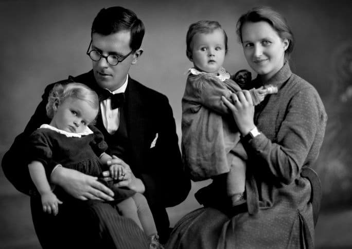 Svartvitt ateljéfotografi av en familj med två vuxna och två små barn. Mannen, klädd i mörk kostym och fluga, sitter med ett barn i knät. Bredvid honom sitter en kvinna i klänning med det andra barnet stående i sitt knä.