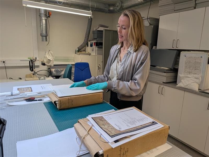 Person med blå handskar står vid bord och rör vid en stor arkivvolym.