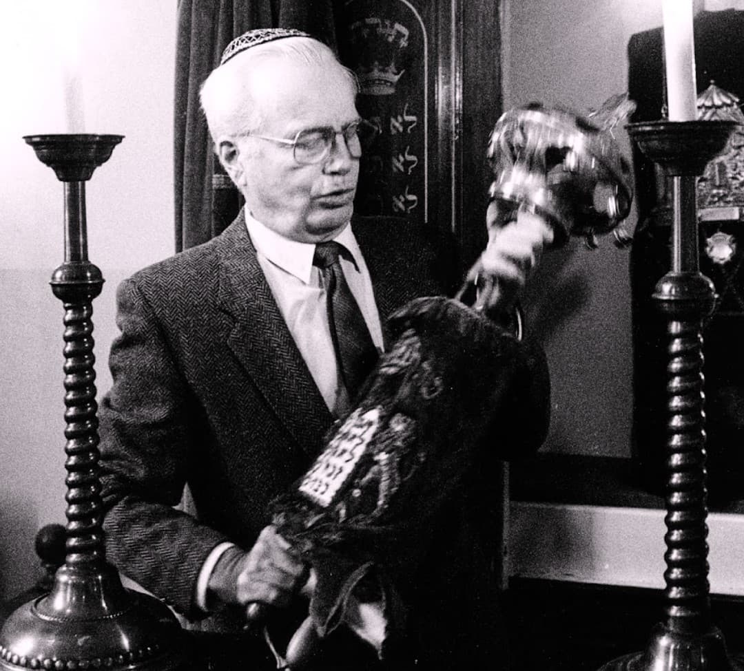 En man med kort vitt hår och rund huvudbonad står mellan två höga ljusstakar. Han håller i ett föremål.