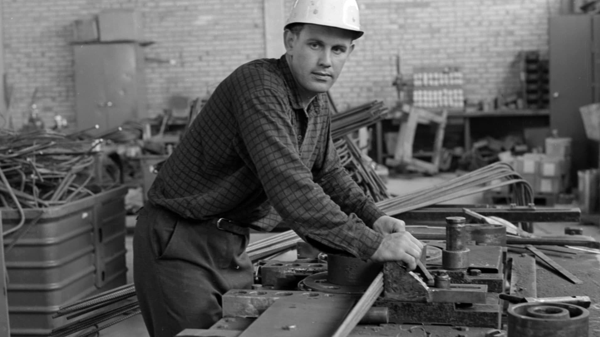 Person i hjälm böjer metallstång med maskin i verkstad.