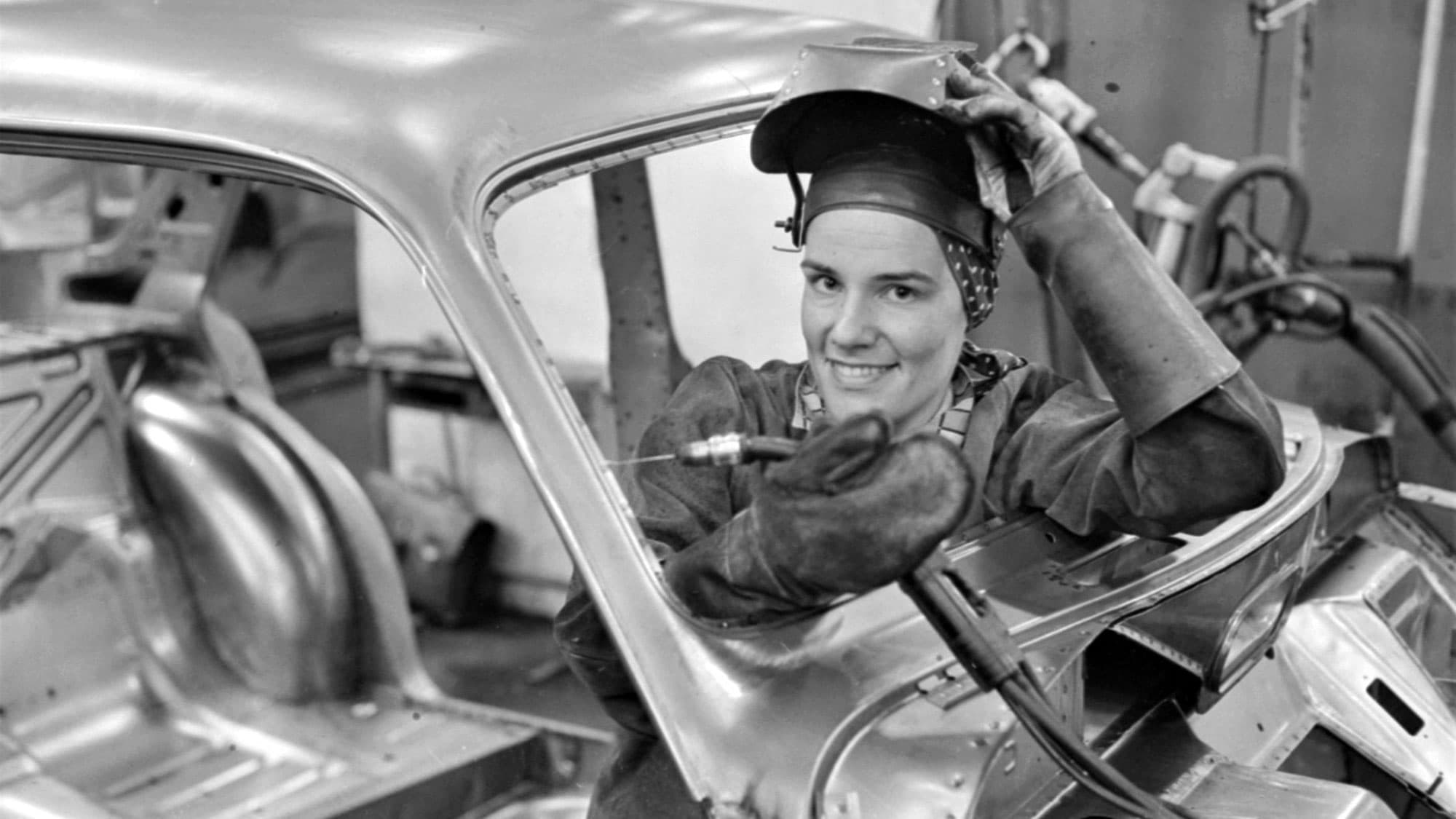 Svetsare ler och lyfter hjälm inne i bilram under arbete.