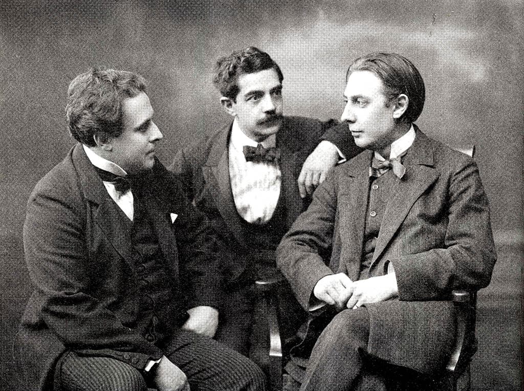 Tre män i 1800-talskläder sitter och samtalar i en studiofotografering.