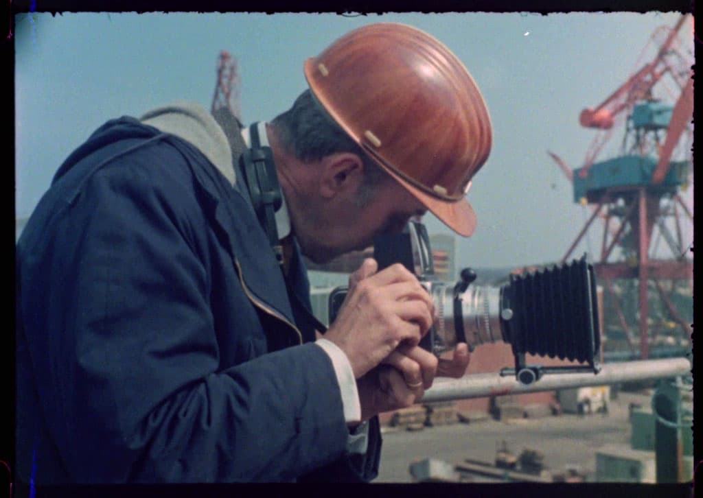 En person med hjälm fotograferar med en kamera på en industriarbetsplats, med kranar i bakgrunden.