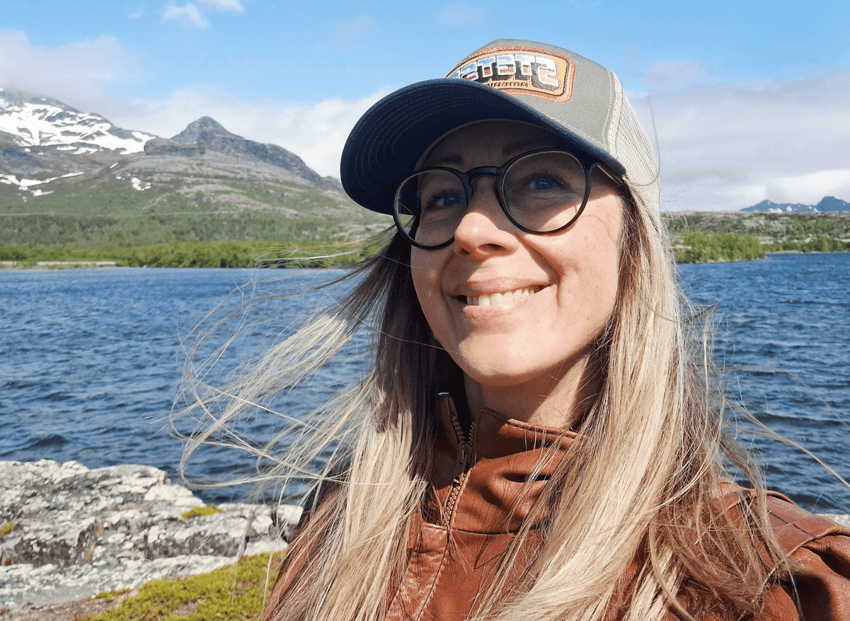 Kvinna med längt hår och keps framför sjö och fjälltopp.